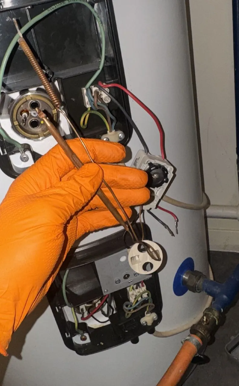 Gloved hand holding an immersion heater thermostat and its copper sensor capillary in front of the open cylinder terminal covers