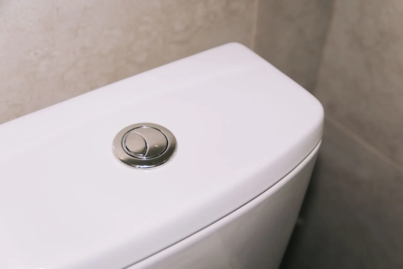 Chrome dual-flush push button on the lid of a white close-coupled toilet cistern against a tiled wall